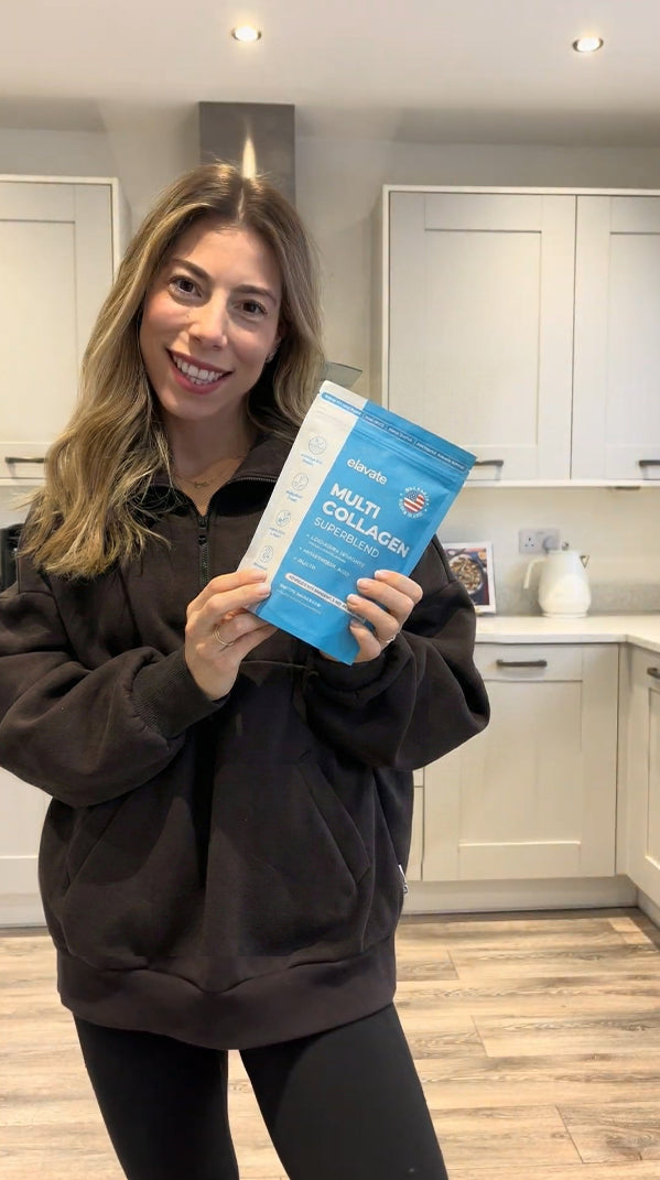Woman in kitchen smiling, holding blue pouch labeled 'elevate MULTI COLLAGEN SUPERBLEND'.